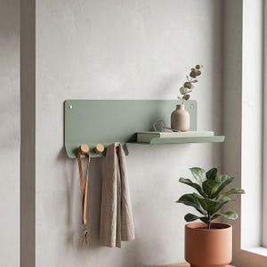 May include: A sage green metal wall shelf with a built-in shelf and two wooden pegs for hanging items. A small vase with greenery, a book, and a pair of glasses are on the shelf. A leather lanyard with keys and a beige scarf hang below.