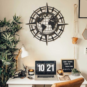 May include: A black metal compass wall decor with a world map design. The compass is hanging on a white wall above a desk with a laptop, a lamp, and a potted plant. The desk also has a letter board with the text "Good things come to those who hustle".