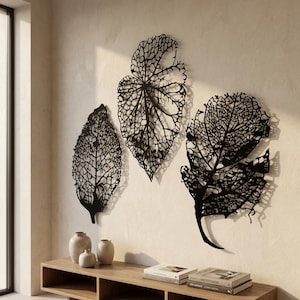 May include: Three black metal leaf wall art pieces with intricate vein detailing are mounted on a light-coloured wall. The leaves vary in size and shape, creating a natural, artistic display. A wooden console with vases and books sits below.