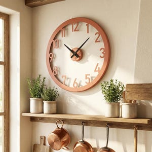Könnte beinhalten: Eine runde Wanduhr mit einem terrakottafarbenen Rahmen und passenden erhabenen Ziffern. Das Zifferblatt ist offen und gibt den Blick auf die Wand frei, mit zwei schwarzen Zeigern, die an einer kleinen terrakottafarbenen Nabe sitzen.