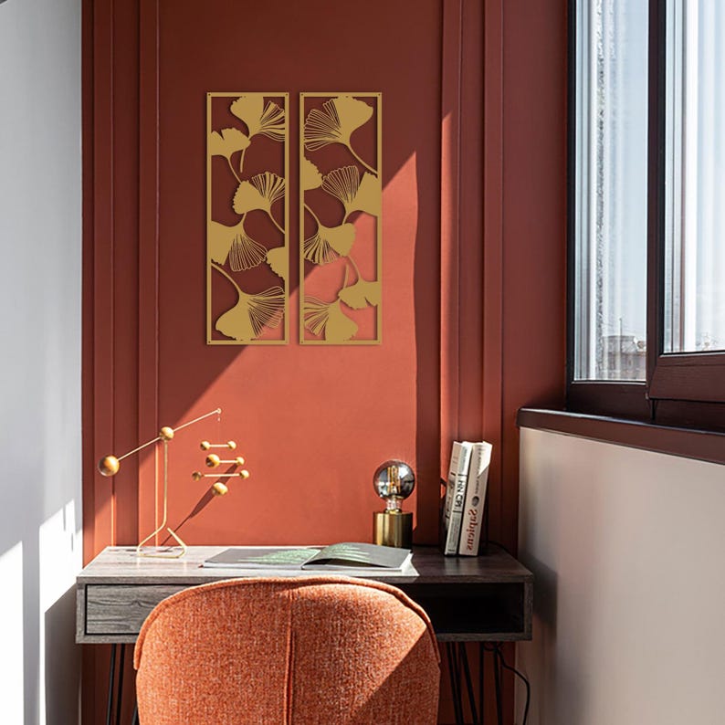 May include: Two gold-colored metal wall art panels with a ginkgo leaf design are mounted on a reddish-brown wall. The panels are rectangular and feature cut-out leaf shapes. A desk with decor and a chair are in front of the wall.