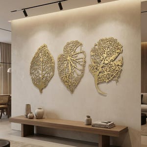 Puede incluir: Tres piezas de arte mural de hojas met&aacute;licas doradas con intrincados cortes en forma de venas. Las hojas est&aacute;n montadas en una pared de color neutro sobre un banco de madera. El arte a&ntilde;ade un elemento decorativo natural a la habitaci&oacute;n.
