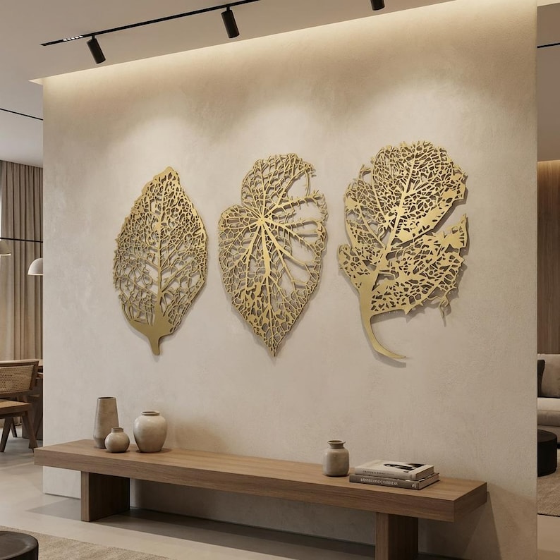 Puede incluir: Tres piezas de arte mural de hojas met&aacute;licas doradas con intrincados cortes en forma de venas. Las hojas est&aacute;n montadas en una pared de color neutro sobre un banco de madera. El arte a&ntilde;ade un elemento decorativo natural a la habitaci&oacute;n.