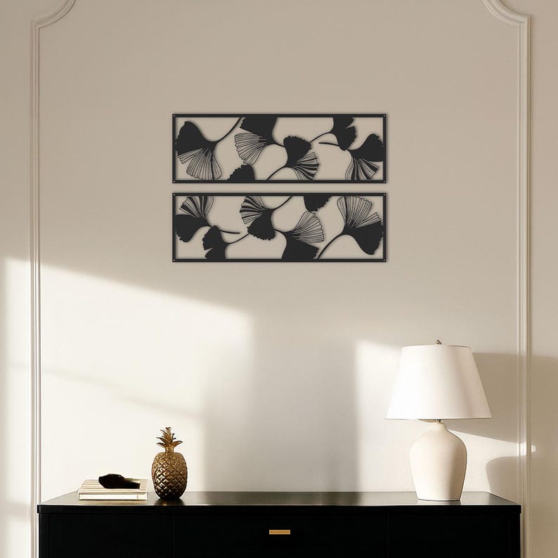 May include: Two rectangular wall art pieces with black ginkgo leaf designs against a cream background. The leaves have detailed vein patterns. The art is framed in black and mounted on a light-colored wall. A black cabinet with a pineapple and lamp are in the foreground.