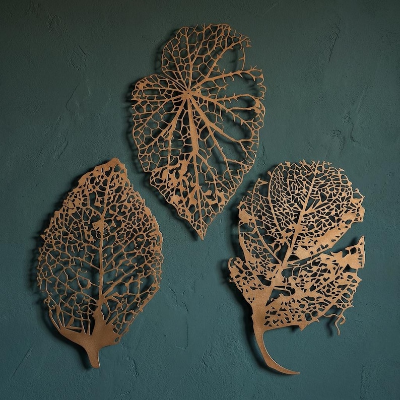 Puede incluir: Tres decoraciones de pared de hojas de metal dorado. Cada hoja presenta un intrincado dise&ntilde;o calado, que muestra las delicadas venas y la estructura de una hoja. Las hojas est&aacute;n montadas en una pared de color verde azulado.