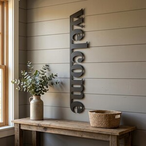 May include: A dark gray metal "welcome" sign is mounted vertically on a gray wall. A wooden table holds a vase with greenery and a woven basket. The scene is lit by natural light from a nearby window.