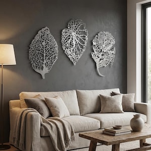 Puede incluir: Tres piezas de arte mural de hojas de metal plateado con intrincados detalles de venas est&aacute;n montadas en una pared gris oscuro. Las hojas son de diferentes formas y tama&ntilde;os. Un sof&aacute; beige con cojines y una mesa de centro de madera est&aacute;n en primer plano.