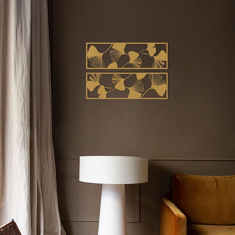 May include: Two rectangular wall art pieces with a brown background and a gold leaf pattern. The leaves are stylized and arranged in a repeating pattern. The art is mounted on a brown wall, with a white lamp and a mustard-colored chair in the foreground.