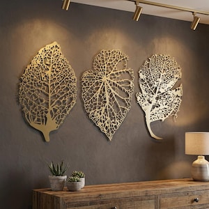 Puede incluir: Tres piezas de arte mural de hojas de metal dorado con intrincados detalles de venas est&aacute;n montadas en una pared gris oscuro. Las hojas tienen diferentes formas y tama&ntilde;os, creando un tema decorativo natural. Debajo se ven un mueble de madera y plantas en macetas.