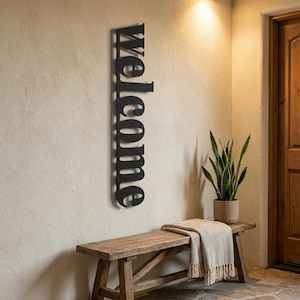 May include: A black metal "Welcome" sign is mounted vertically on a beige wall. The sign is in a bold, sans-serif font. A wooden bench with a beige throw blanket and a potted plant are below the sign. The scene is well-lit, suggesting an entryway.
