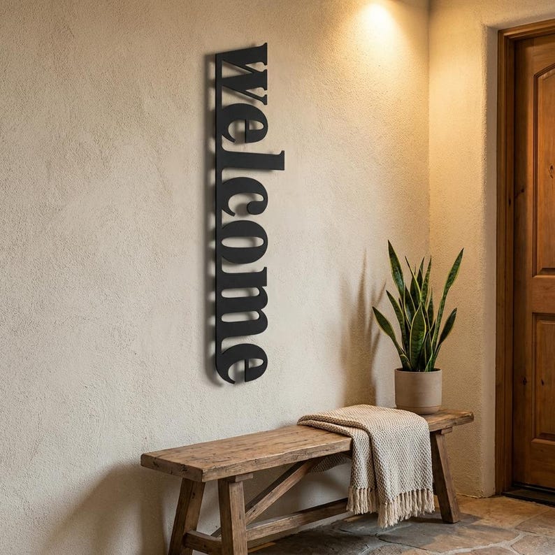 May include: A black metal "Welcome" sign is mounted vertically on a beige wall. The sign is in a bold, sans-serif font. A wooden bench with a beige throw blanket and a potted plant are below the sign. The scene is well-lit, suggesting an entryway.
