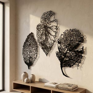 Puede incluir: Tres piezas de arte mural de hojas de metal negro con intrincados patrones de venas est&aacute;n montadas en una pared de color claro. Las hojas var&iacute;an en tama&ntilde;o y forma, creando una est&eacute;tica natural y org&aacute;nica. Debajo hay una consola de madera con jarrones y libros.