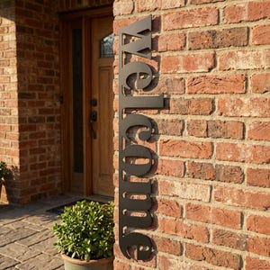 May include: A black metal "welcome" sign is mounted on a red brick wall. The sign is vertically oriented and features a cursive font. A wooden door and potted plant are visible in the background.