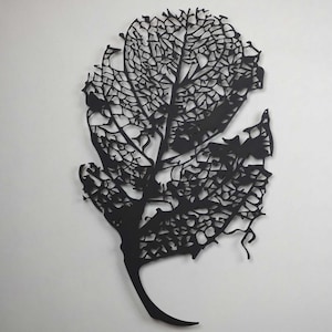 Puede incluir: Arte de pared de hoja de metal negro. El intrincado dise&ntilde;o presenta una estructura esquel&eacute;tica detallada de hoja, mostrando las venas y los bordes delicados. La obra de arte est&aacute; montada en una pared blanca.