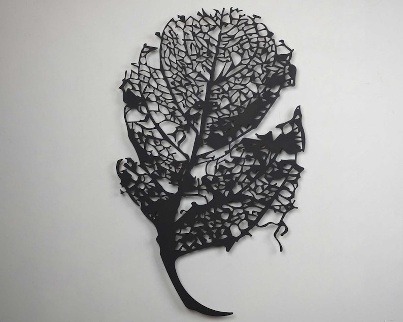 Puede incluir: Arte de pared de hoja de metal negro. El intrincado dise&ntilde;o presenta una estructura esquel&eacute;tica detallada de hoja, mostrando las venas y los bordes delicados. La obra de arte est&aacute; montada en una pared blanca.