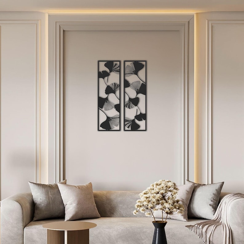 May include: Two black metal wall art panels, each with a rectangular frame, feature a ginkgo leaf design. The leaves are black with intricate vein details, set against a neutral background. The artwork is displayed above a beige sofa.