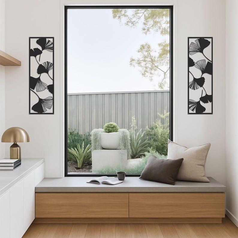 May include: Two black metal wall art pieces with a ginkgo leaf design, flanking a large window. The leaves are black against a cream background. The room features a built-in bench with pillows and a view of a garden.