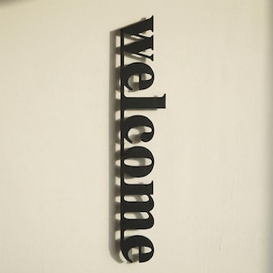 May include: A black metal sign that reads "welcome" in a vertical orientation. The sign is mounted on a white wall. The letters are in a bold, sans-serif font.