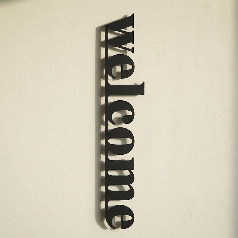 May include: A black metal sign that reads "welcome" in a vertical orientation. The sign is mounted on a white wall. The letters are in a bold, sans-serif font.