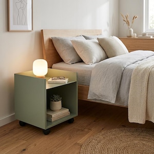 May include: A sage green bedside table with a glowing white lamp, books, and a small potted plant. The table has a shelf and sits on small black wheels. The bed has light gray bedding and a beige throw.