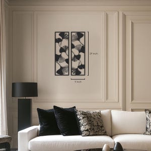 May include: Two black metal wall art panels, each 9 inches wide and 29 inches tall, featuring a ginkgo leaf design. The panels are displayed on a beige wall above a white sofa with black pillows.