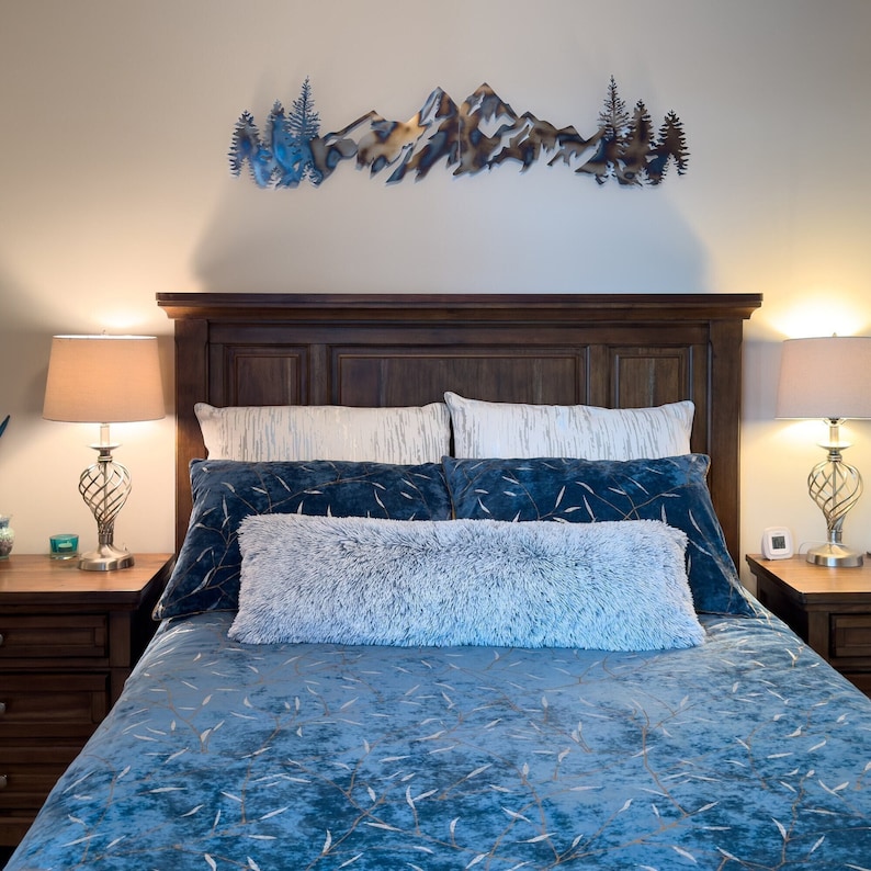 May include: A blue and white patterned bedspread with a fluffy white throw pillow on a bed with a dark wood headboard. A metal mountain range wall art hangs above the bed.