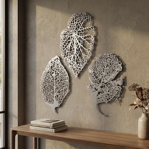 Puede incluir: Tres piezas de arte mural de hojas de metal plateado con intrincados patrones de venas est&aacute;n montadas en una pared de tono neutro. Las hojas son de varios tama&ntilde;os y formas, creando una est&eacute;tica natural y org&aacute;nica. Una consola de madera con libros y un jarr&oacute;n de flores secas est&aacute; en primer plano.