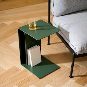 May include: A modern, dark green side table with a built-in shelf holding two white books. A clear glass with a yellow liquid sits on the tabletop. The table is next to a gray upholstered chair.