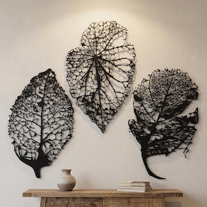 Puede incluir: Tres obras de arte murales de hojas de metal negro con intrincados patrones de venas est&aacute;n montadas en una pared blanca. Las hojas tienen diferentes formas y tama&ntilde;os, creando una exhibici&oacute;n art&iacute;stica natural. Un peque&ntilde;o jarr&oacute;n y libros descansan sobre una mesa de madera.