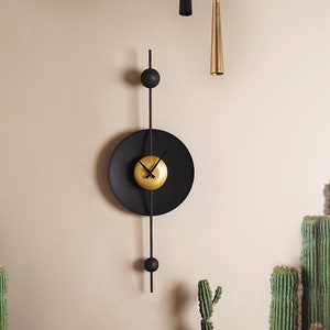 May include: A contemporary wall clock featuring a black circular face with gold hands and centre. The clock has a minimalist design with a black vertical rod and two black spheres. It is mounted on a beige wall.