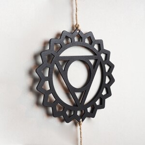 Op de afbeelding: Zwarte houten mandala met een geometrisch ontwerp, hangend aan een touwtje van touw. De mandala is een symbool van de vijfde chakra, de keelchakra.