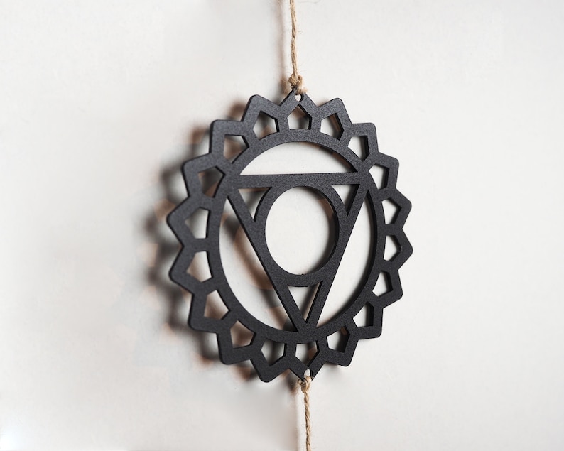 Op de afbeelding: Zwarte houten mandala met een geometrisch ontwerp, hangend aan een touwtje van touw. De mandala is een symbool van de vijfde chakra, de keelchakra.