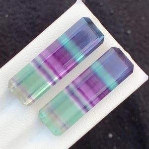 May include: Two rectangular fluorite gemstones. Each stone features bands of purple, blue, and green. The gemstones are displayed on a white surface against a dark background. The stones have a polished, translucent appearance.