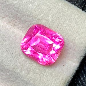 May include: A vivid, cushion-cut, hot pink gemstone. The faceted gem is set against a soft, off-white fabric, with a dark, textured background. The gemstone's cut creates a brilliant display of light and colour.