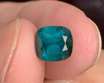 Può includere: Una pietra preziosa verde scuro con taglio a cuscino. La pietra preziosa è tenuta in una mano.