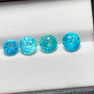 May include: Four round, faceted gemstones in a light blue to teal colour. The gemstones are arranged in a row on a white surface.