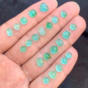 May include: A hand holds a selection of small, polished emerald gemstones. The gems are in various shapes, including round, square, and oval, and are a vibrant green colour. The gemstones are of various sizes, with the largest being approximately 1.3 cm in diameter.