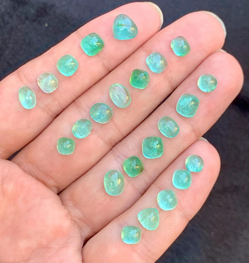 May include: A hand holds a selection of small, polished emerald gemstones. The gems are in various shapes, including round, square, and oval, and are a vibrant green colour. The gemstones are of various sizes, with the largest being approximately 1.3 cm in diameter.