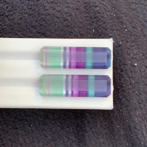 May include: Two rectangular fluorite gemstones with bands of green, purple, and blue. The stones are set on a white surface against a dark, textured background. The gemstones have a polished, faceted appearance. The gemstones are likely for jewellery.