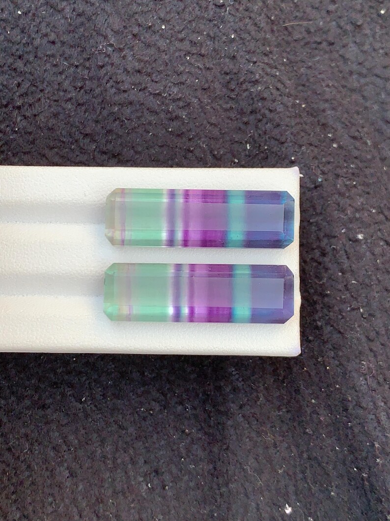 May include: Two rectangular fluorite gemstones with bands of green, purple, and blue. The stones are set on a white surface against a dark, textured background. The gemstones have a polished, faceted appearance. The gemstones are likely for jewellery.