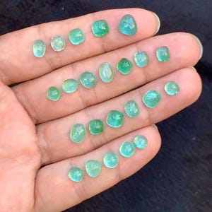 May include: A selection of small, polished gemstones in various shapes and sizes, held in a hand. The gems display a range of green and teal hues, with a translucent quality. The arrangement showcases the variety of cuts and colours.