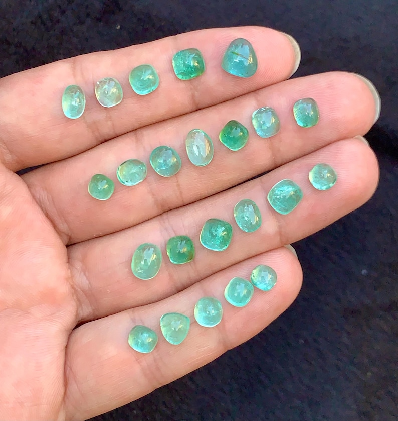 May include: A selection of small, polished gemstones in various shapes and sizes, held in a hand. The gems display a range of green and teal hues, with a translucent quality. The arrangement showcases the variety of cuts and colours.