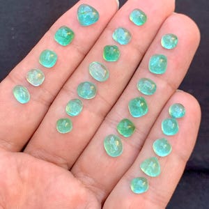 May include: A selection of small, polished gemstones in various shapes, including round, oval, and square. The gems are a vibrant teal colour, with a translucent quality. The stones are displayed on a light-toned surface.