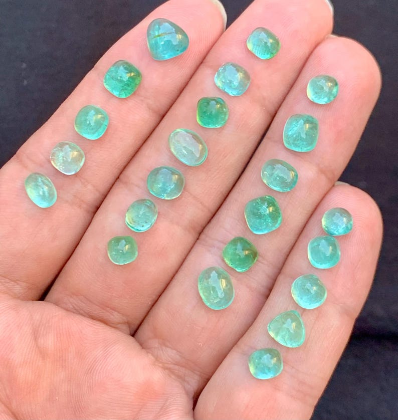 May include: A selection of small, polished gemstones in various shapes, including round, oval, and square. The gems are a vibrant teal colour, with a translucent quality. The stones are displayed on a light-toned surface.