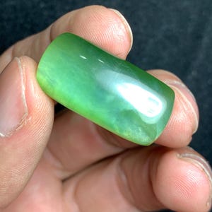 Può includere: Una gemma rettangolare lucidata con una vibrante tonalità verde. La pietra ha bordi arrotondati e una superficie liscia e riflettente. La gemma è tenuta nella mano di una persona.
