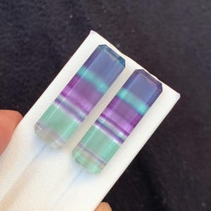 May include: Two rectangular fluorite crystals with a gradient of colours, including purple, green, and blue. The crystals are displayed on a white surface, showcasing their polished, translucent appearance. The crystals are held by a hand.