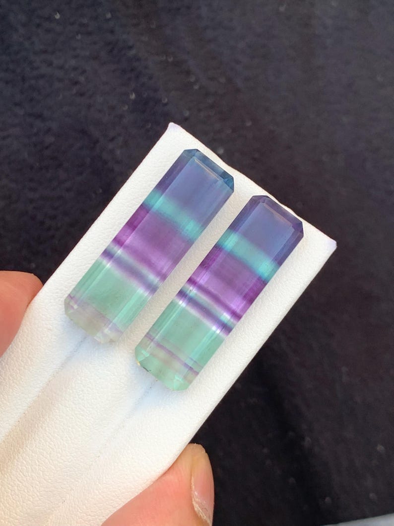 May include: Two rectangular fluorite crystals with a gradient of colours, including purple, green, and blue. The crystals are displayed on a white surface, showcasing their polished, translucent appearance. The crystals are held by a hand.