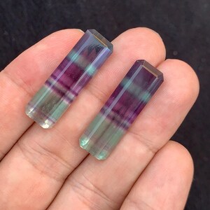 May include: Two rectangular fluorite crystals. Each crystal is a mix of purple, green, and clear colours. The crystals are held in a hand against a dark background. The crystals have a polished, faceted appearance.