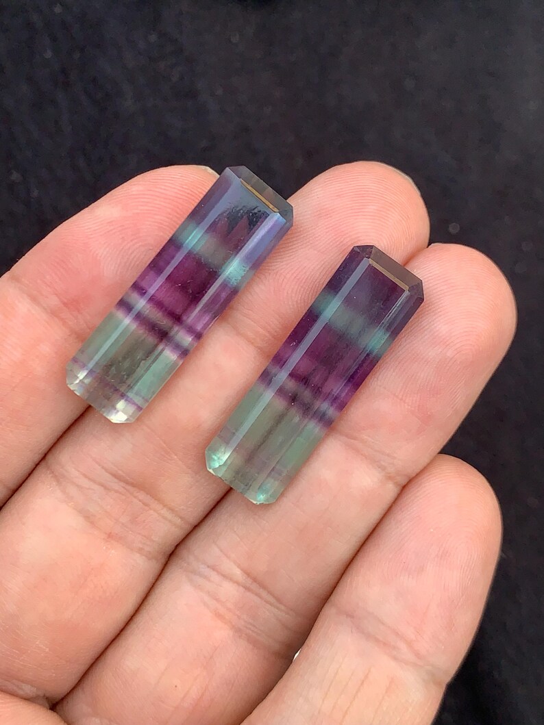May include: Two rectangular fluorite crystals. Each crystal is a mix of purple, green, and clear colours. The crystals are held in a hand against a dark background. The crystals have a polished, faceted appearance.