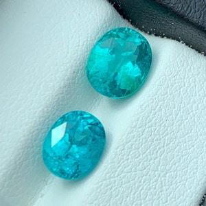 May include: Two oval-shaped turquoise gemstones on a white background. The gemstones are faceted and have a bright, vibrant colour.